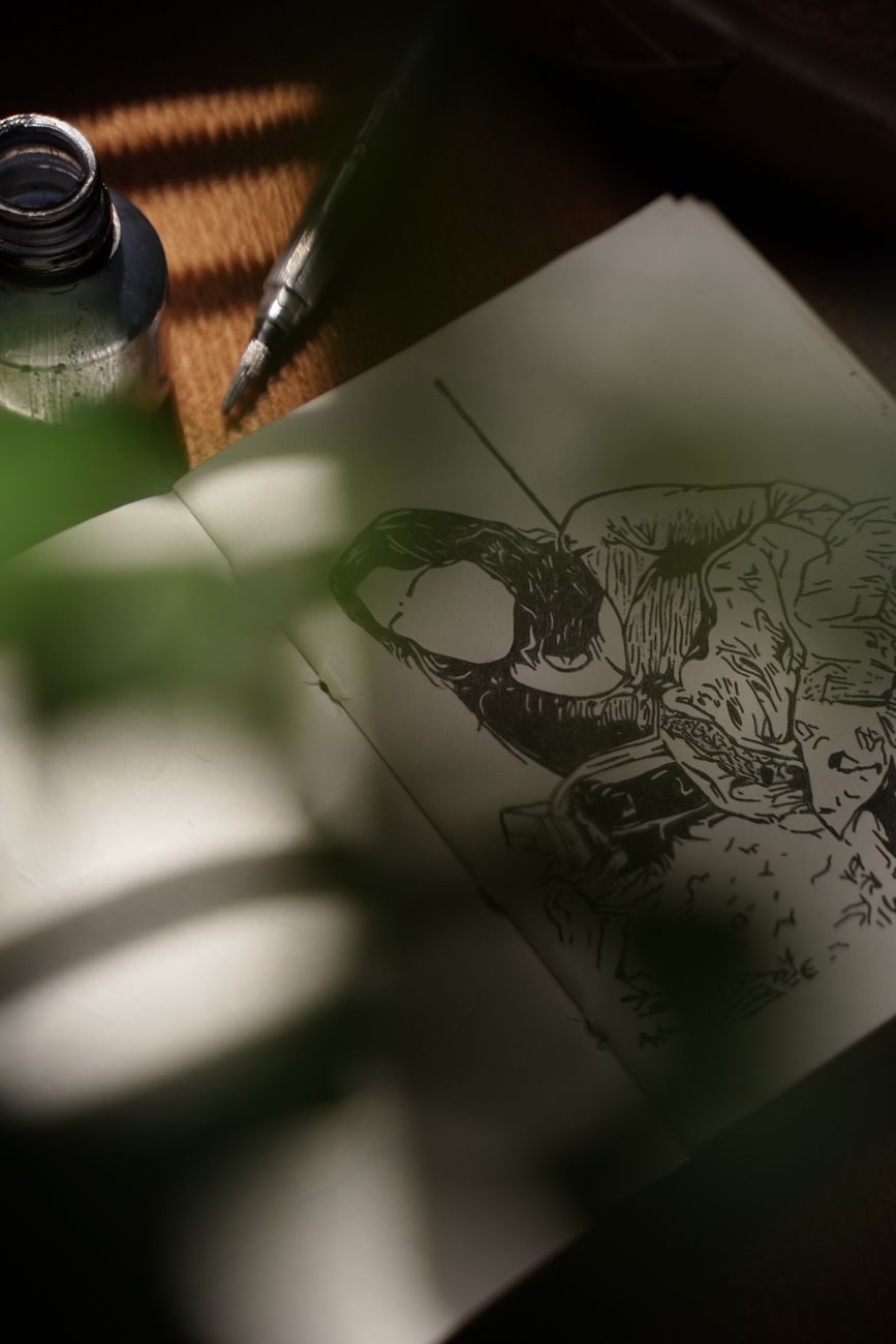 An open sketchbook with an ink drawing of a figure wearing a headset, lying next to a bottle of ink and a dip pen on a wooden table.