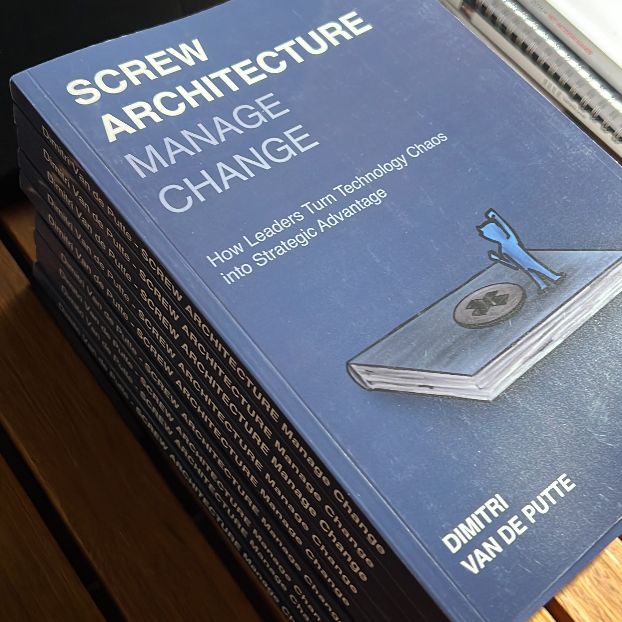 A stack of copies of the book "Screw Architecture, Manage Change" by Dimitri Van de Putte lies on a wooden table.