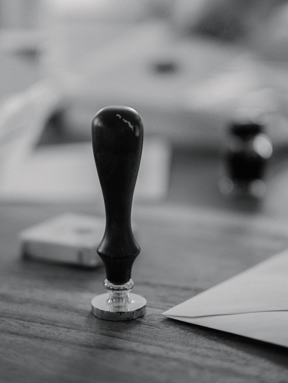 A black-and-white close-up of a wax stamp standing upright on a wooden desk next to an envelope and an ink pad.